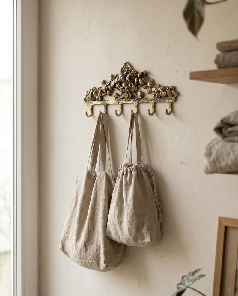May include: A decorative brass wall hook with floral detailing, holding two beige linen bags. The hook has five curved hooks for hanging items. The bags are different sizes and have a natural, neutral color.