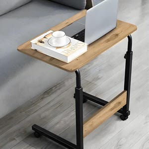 May include: A mobile laptop desk with a light brown wooden surface and black metal frame. A laptop, a book, and a teacup and saucer are on the desk. The desk has wheels for easy movement and is adjustable in height.
