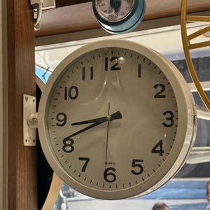 May include: A round, white wall-mounted quartz clock with a clean white face, black Arabic numerals, and black hour, minute, and second hands. The clock is attached to a wooden frame by a white metal bracket, featuring a simple, functional design.