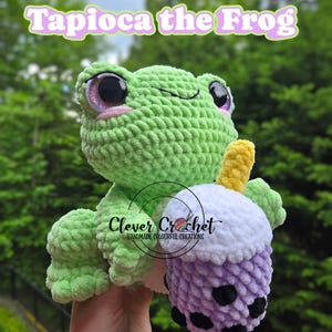 Puede incluir: Un peluche de rana verde tejido a ganchillo que sostiene un bubble tea morado y blanco tejido a ganchillo. La rana tiene ojos grandes y brillantes y el texto "Tapioca the Frog" está encima. El bubble tea tiene bobas negras y una pajita amarilla.