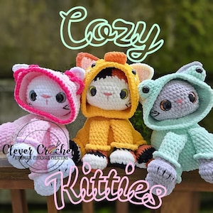 Op de afbeelding: Drie handgemaakte gehaakte kattenpoppen in roze, geel en blauw, elk met een outfit met capuchon. Het woord "Cozy" staat boven de poppen en het woord "Kitties" eronder. De poppen zitten op een houten oppervlak.