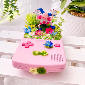 May include: A pink Nintendo Game Boy decorated with a colorful floral arrangement and a Mr. Mime figurine. The handheld console features a D-pad, buttons, and speaker holes. The floral arrangement includes blue, pink, and yellow flowers, and green foliage.