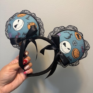 May include: A pair of black headband ears with a Halloween theme. The round teal ears feature Jack Skellington faces, pumpkins, and sweets. Black lace trim and black bows complete the design.