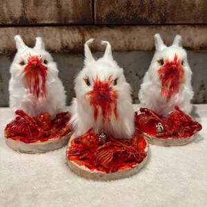 May include: Three white faux fur rabbit figurines with red painted faces and chests, each mounted on a circular wooden base. The bases are covered in red textured debris, including small skeletal remains and a miniature silver grenade charm.