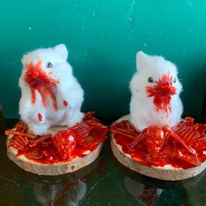 May include: Two white plush bunny figurines with red blood-like paint on their faces and surrounding a wooden base. The bases feature a red skull and bone design, creating a spooky, Halloween-themed decoration.
