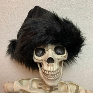 May include: A black faux fur hat with a black felt crown. The hat is worn by a white plastic skeleton.