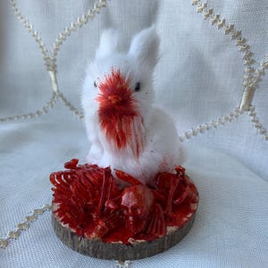 May include: A small white fuzzy rabbit figurine with red paint on its face and chest, positioned on a circular wooden base covered in red-painted skeletal remains and a skull, creating a stark contrast between the white fur and red details.