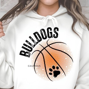 May include: White hoodie featuring the word "BULLDOGS" in black above an orange basketball graphic with black lines and a black paw print. The word "CAUTIOUS" is printed below the basketball.