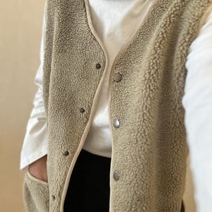 May include: A long, beige fleece vest with a button closure and two pockets. The vest has a brown trim around the edges.