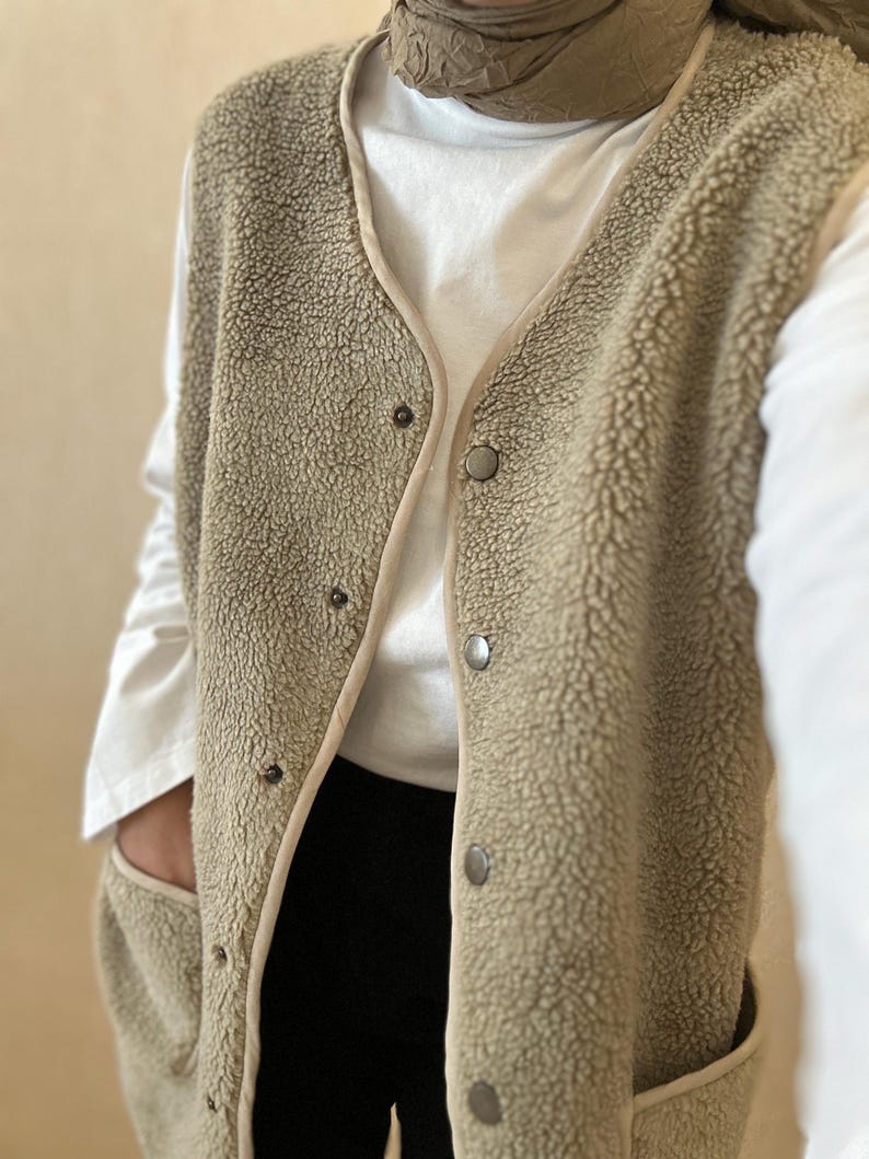 May include: A long, beige fleece vest with a button closure and two pockets. The vest has a brown trim around the edges.