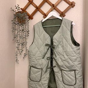 May include: A light green quilted vest with a tie closure and two patch pockets. The vest is hanging on a wooden rack.