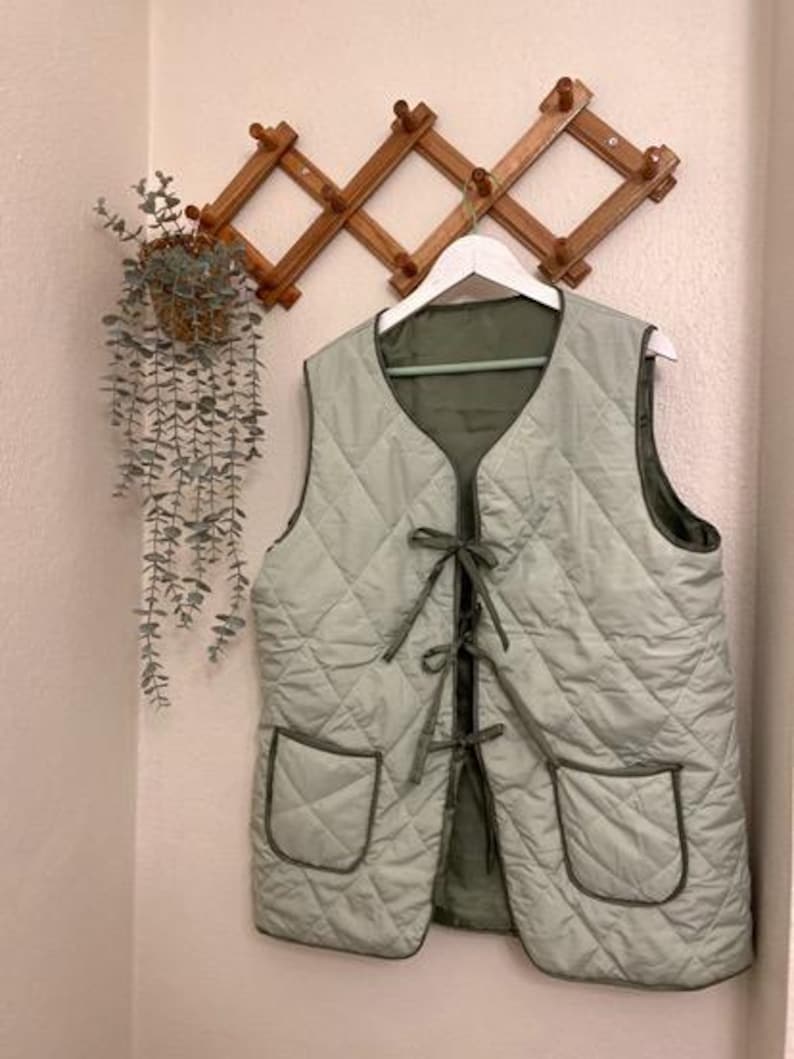 May include: A light green quilted vest with a tie closure and two patch pockets. The vest is hanging on a wooden rack.