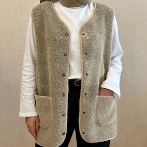 May include: A beige sherpa vest with a button closure and two large pockets. The vest has a brown trim around the edges and pockets.