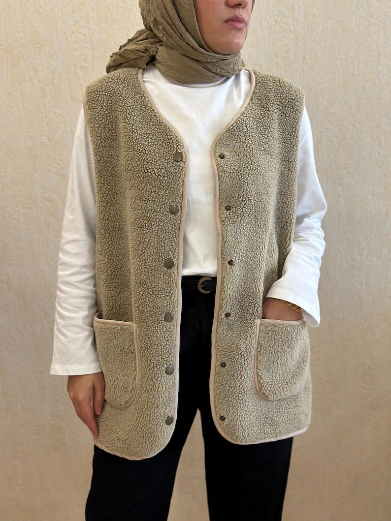 May include: A beige sherpa vest with a button closure and two large pockets. The vest has a brown trim around the edges and pockets.