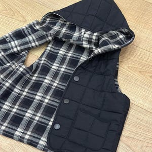 May include: Black quilted vest with a plaid flannel lining and a hood. The vest has a zip fastening and two button fastenings.