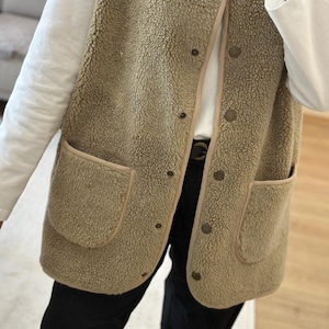 May include: A beige fleece vest with a button closure and two pockets. The vest has a brown trim around the edges and pockets.