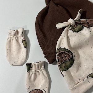 May include: A set of baby accessories featuring a brown knit hat, a white and brown patterned bib, and a pair of white and brown patterned mittens with a cartoon chestnut character wearing glasses.