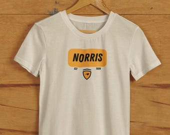May include: Off-white t-shirt featuring the word "NORRIS" in a yellow rectangle. Below is a shield logo and the text "EST 1999". The t-shirt is displayed on a wooden hanger.