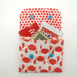 May include: A small, open wallet with a red crab pattern on a white background. The interior contains a red card, a dollar bill, and a packet. The top flap has a red polka dot pattern and is secured with two blue snaps.