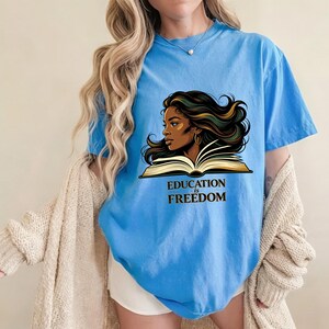 May include: A light blue short-sleeved t-shirt featuring a graphic illustration of a person with long flowing dark hair reading an open book. Below the illustration, the text EDUCATION IS FREEDOM is printed in a bold, dark serif font.