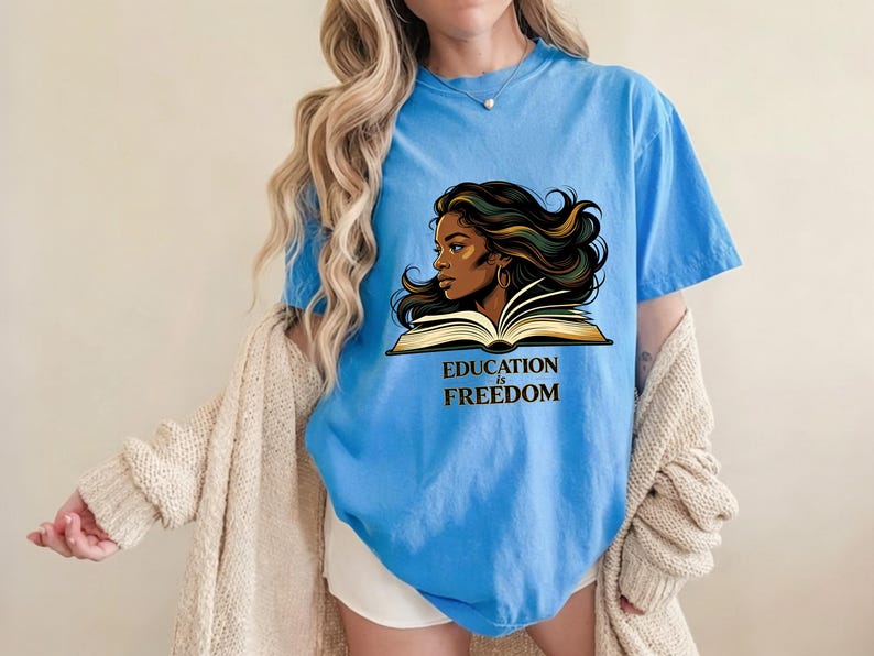 May include: A light blue short-sleeved t-shirt featuring a graphic illustration of a person with long flowing dark hair reading an open book. Below the illustration, the text EDUCATION IS FREEDOM is printed in a bold, dark serif font.