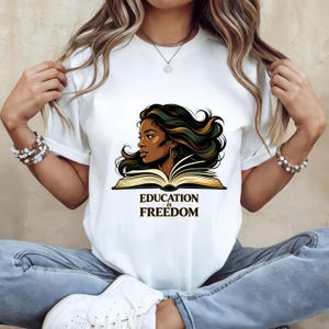 May include: A white short-sleeved t-shirt featuring a graphic illustration of a person with long dark hair looking over an open book. Below the illustration, the text reads "EDUCATION is FREEDOM" in a bold, dark, serif font style.