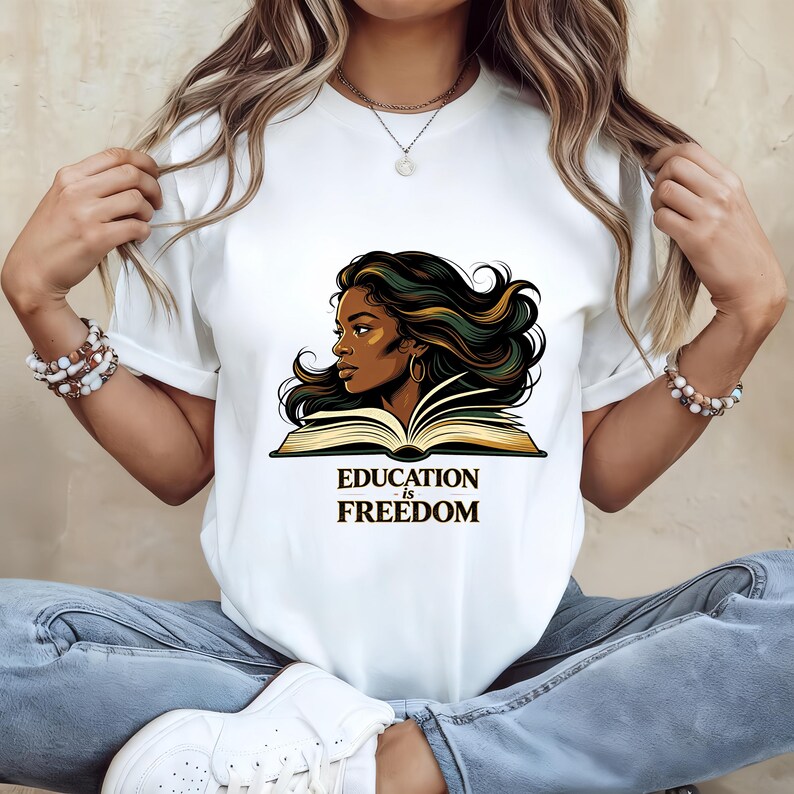 May include: A white short-sleeved t-shirt featuring a graphic illustration of a person with long dark hair looking over an open book. Below the illustration, the text reads "EDUCATION is FREEDOM" in a bold, dark, serif font style.