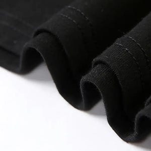 Op de afbeelding: Close-up van zwarte stof met zichtbare stiksels. De stof is gevouwen, waardoor zachte rondingen en schaduwen ontstaan. De textuur van het materiaal is duidelijk zichtbaar, waarbij de stiksels details aan het kledingstuk toevoegen.
