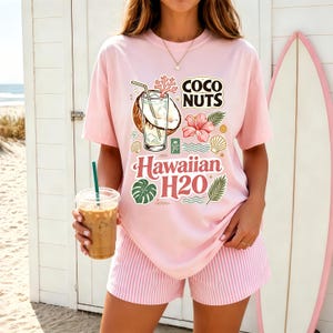 May include: A light pink t-shirt featuring a graphic print of a coconut drink, a pink hibiscus flower, and tropical leaves. The text reads COCO NUTS and Hawaiian H2O. The shirt is paired with light pink and white vertically striped shorts.