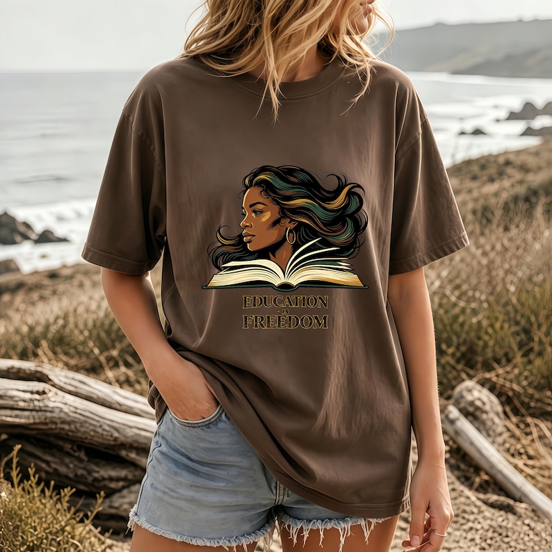 May include: A brown short-sleeve t-shirt featuring a graphic illustration of a person with long flowing hair positioned behind an open book. Below the book, the text reads "EDUCATION IS FREEDOM" in a dark, stylised serif font.