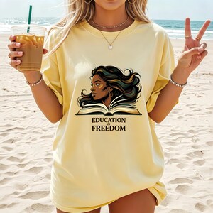 May include: A pale yellow short-sleeve t-shirt featuring a graphic illustration of a person with long dark hair reading an open book. Below the illustration, the text reads "EDUCATION is FREEDOM" in a bold, dark serif font centred on the chest.