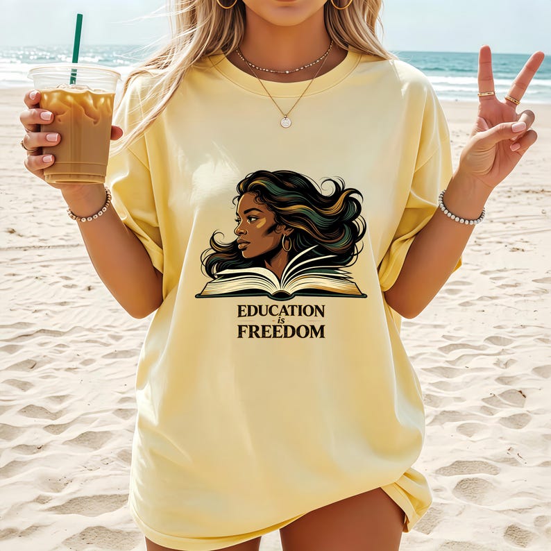 May include: A pale yellow short-sleeve t-shirt featuring a graphic illustration of a person with long dark hair reading an open book. Below the illustration, the text reads "EDUCATION is FREEDOM" in a bold, dark serif font centred on the chest.