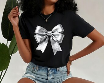 May include: Black t-shirt featuring a large, metallic silver bow graphic. The bow has a textured, shiny appearance. The shirt is a classic crew neck style and is paired with denim shorts. The overall look is casual and festive.