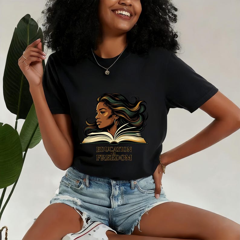 May include: A black short-sleeved t-shirt featuring a graphic illustration of a person with long flowing hair looking over an open book. Below the book, the text reads EDUCATION IS FREEDOM in a gold-coloured serif font.
