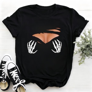 Op de afbeelding: Zwart T-shirt met een grafisch ontwerp van een gescheurde borst, met een huidkleurig gebied en witte skelet handen. Zwarte en witte canvas sneakers completeren de look, wat wijst op een casual Halloween-stijl.