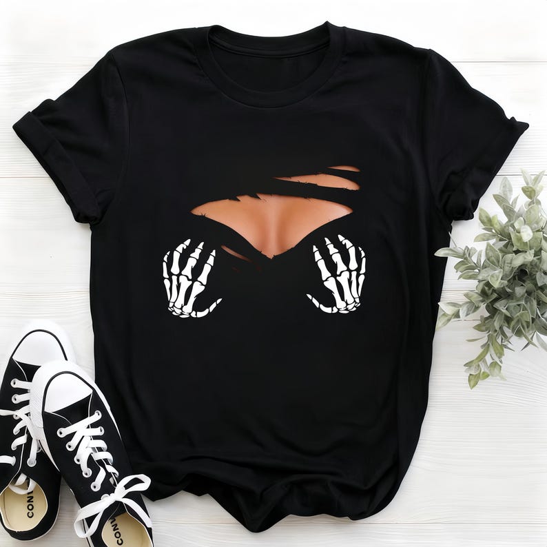 Op de afbeelding: Zwart T-shirt met een grafisch ontwerp van een gescheurde borst, met een huidkleurig gebied en witte skelet handen. Zwarte en witte canvas sneakers completeren de look, wat wijst op een casual Halloween-stijl.