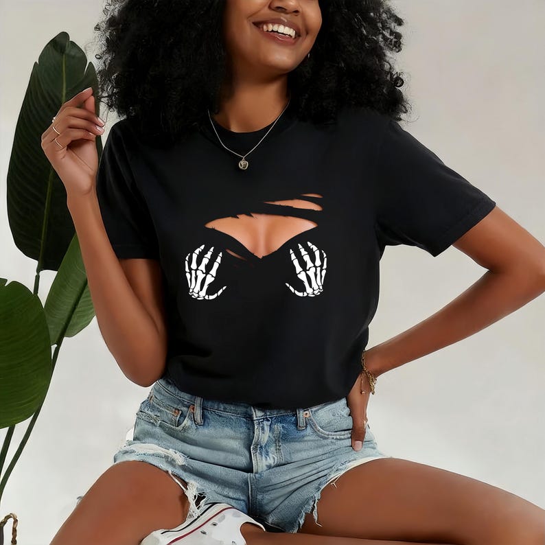 Op de afbeelding: Zwart T-shirt met een gescheurd ontwerp dat een borst en witte skelet handen onthult. Het shirt heeft korte mouwen en een ronde hals. De persoon draagt een spijkerbroek short.