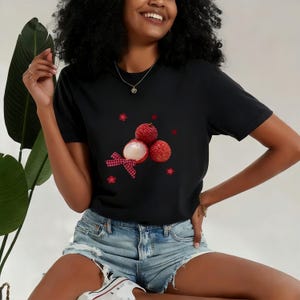 May include: Black t-shirt featuring a vibrant graphic of lychees with a red and white gingham bow. The design includes red flower and star accents. The shirt is paired with denim shorts.