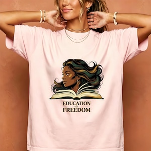 May include: A light pink t-shirt featuring a graphic illustration of a person with dark flowing hair positioned behind an open book. Below the illustration, the text reads "EDUCATION is FREEDOM" in a bold, dark serif font.