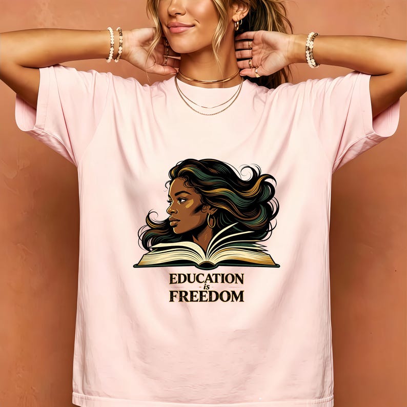 May include: A light pink t-shirt featuring a graphic illustration of a person with dark flowing hair positioned behind an open book. Below the illustration, the text reads "EDUCATION is FREEDOM" in a bold, dark serif font.