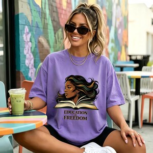 May include: A lavender short-sleeved t-shirt featuring a graphic illustration of a person with long dark hair reading an open book. Below the illustration, the text reads "EDUCATION is FREEDOM" in a dark brown serif font. The shirt has a crew neckline.