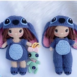 Puede incluir: Dos muñecas de ganchillo hechas a mano, una con un vestido azul y la otra con un mono azul, ambas con sombreros azules a juego con orejas grandes y ojos negros. También hay una pequeña muñeca verde con un lazo rosa. Las muñecas están hechas con costuras detalladas.
