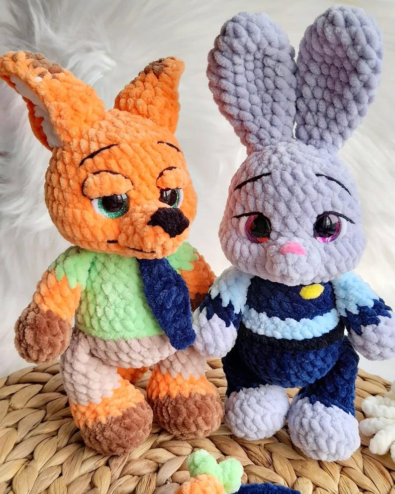 May include: Two handmade crochet toys: an orange fox with green and blue details and a grey bunny with blue and white accents. Both have detailed faces and are made with soft, textured yarn. The toys are displayed on a woven surface.