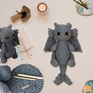 May include: Two handmade gray crochet dragon toys with yellow eyes and wings. One dragon is standing upright, and the other is sitting. The image also shows crochet hooks, yarn, and a small candle.
