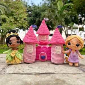 May include: A collection of crocheted princess dolls and a pink castle. The dolls include a princess in a green and yellow dress and another in a purple dress. The castle has pink turrets and a pink and purple facade.