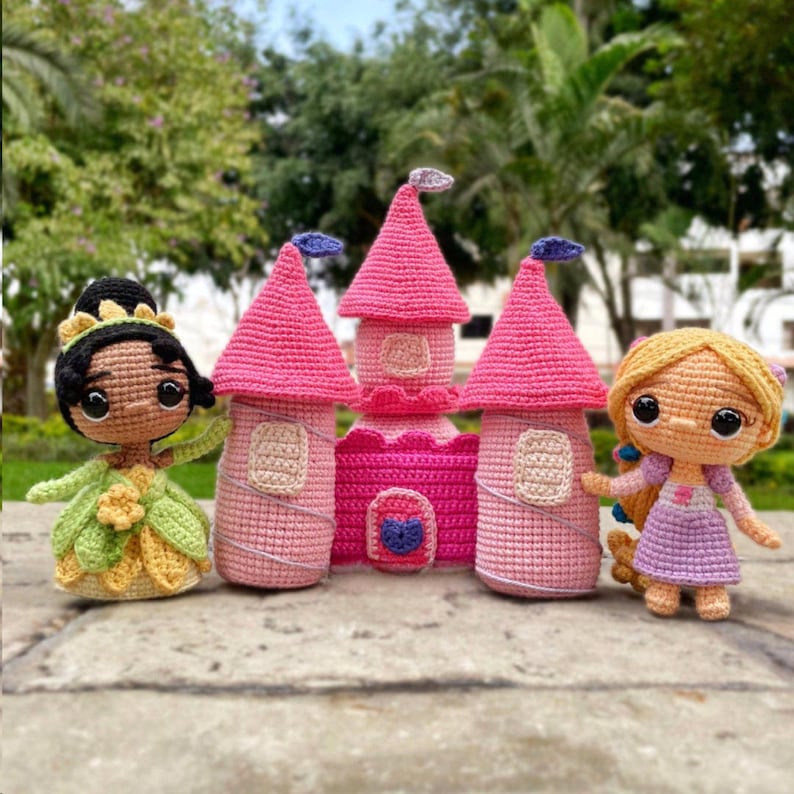May include: A collection of crocheted princess dolls and a pink castle. The dolls include a princess in a green and yellow dress and another in a purple dress. The castle has pink turrets and a pink and purple facade.