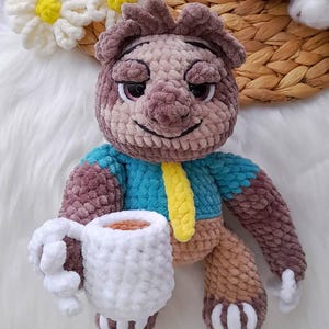 May include: A crocheted bear doll with a blue top and yellow tie, holding a white mug. The bear has brown and tan fur, with a smiling face. The mug appears to contain a brown liquid. The doll is on a white surface.