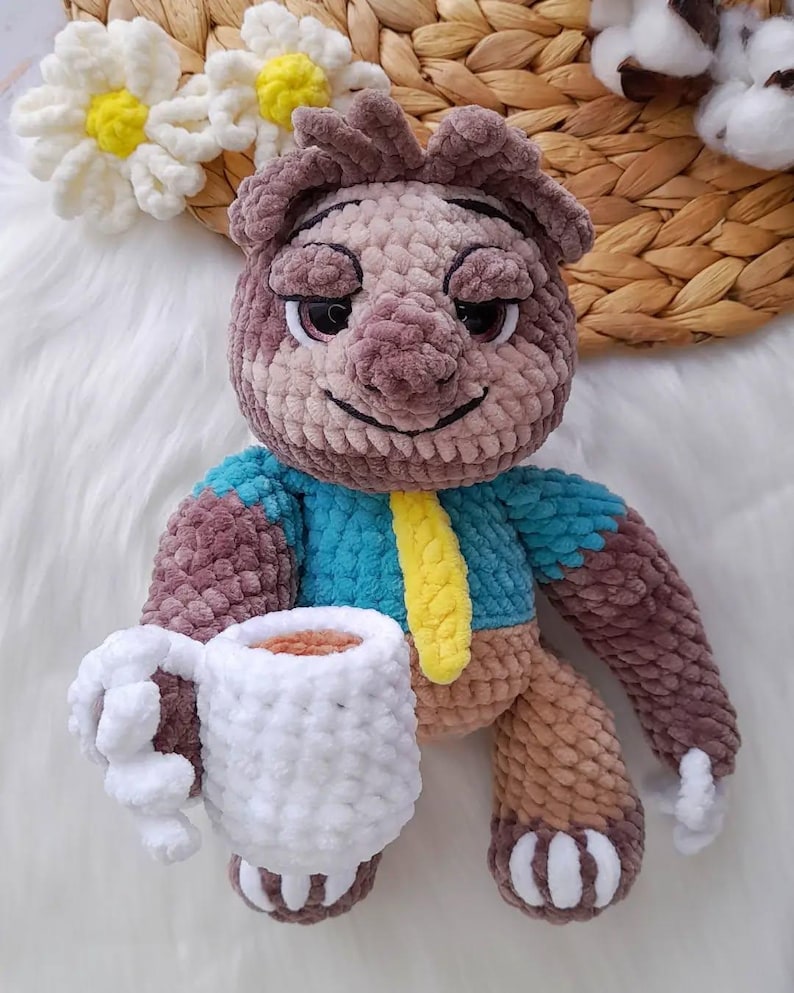 May include: A crocheted bear doll with a blue top and yellow tie, holding a white mug. The bear has brown and tan fur, with a smiling face. The mug appears to contain a brown liquid. The doll is on a white surface.