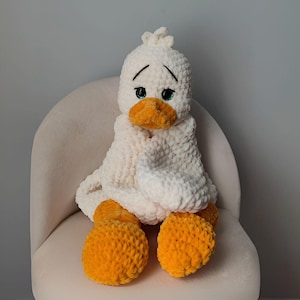May include: A handmade, plush duck toy with a sad expression. The duck is primarily white with a fluffy texture, featuring orange feet and beak. It has black eyebrows and teal-colored eyes. The toy is sitting on a cream-colored chair.