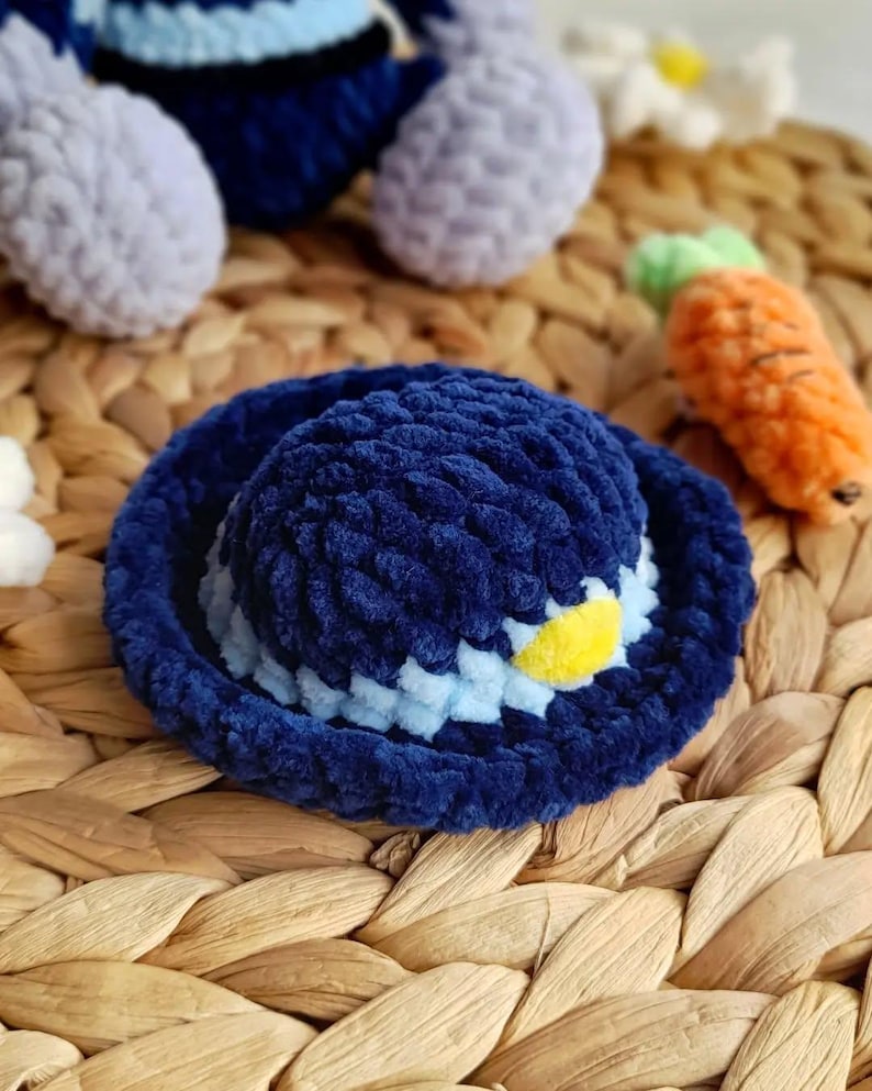 May include: A navy blue crocheted hat with a light blue and yellow accent. The hat is made of a soft, plush material and has a rounded crown and a wide brim. The hat is part of a set of crocheted toys.